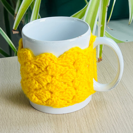 White mug with a yellow knitted cozy on a wooden surface with plants in the background