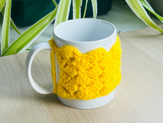 White mug with a yellow knitted cozy on a wooden surface, with plants in the background.